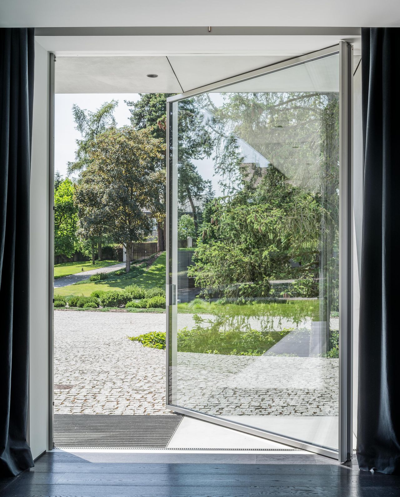 Full-height Sky-Frame Pivot glass door standing open from a dark interior onto a cobbled drive and garden
