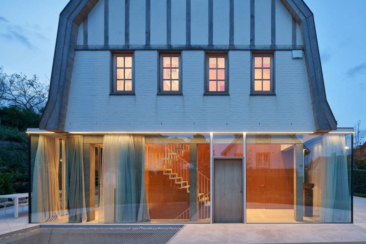 Gabled house at dusk, its ground floor glazed end to end with Sky-Frame behind sheer curtains